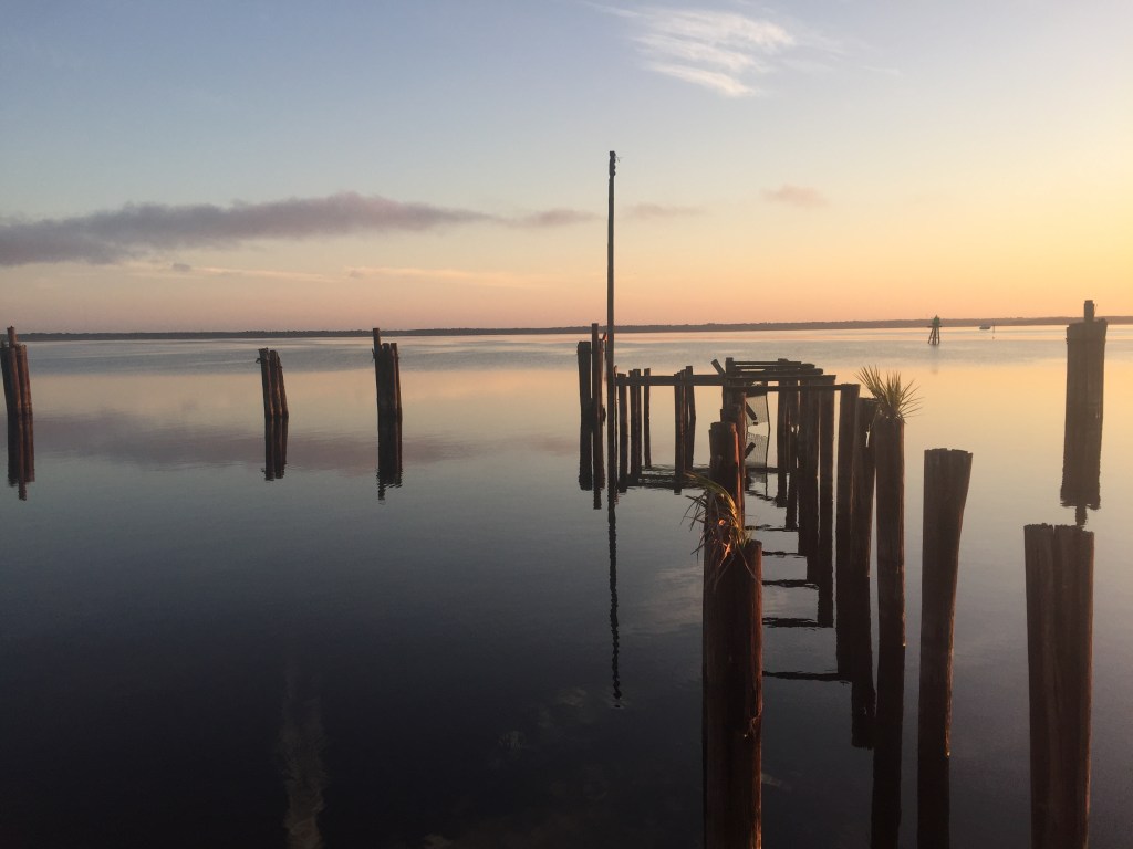lake with still water and posts in a geometric arrangement. Color palette is soft pastelle, with lake water darker in the foreground and turning lighter as the water meets the sky on the horizon in the background.